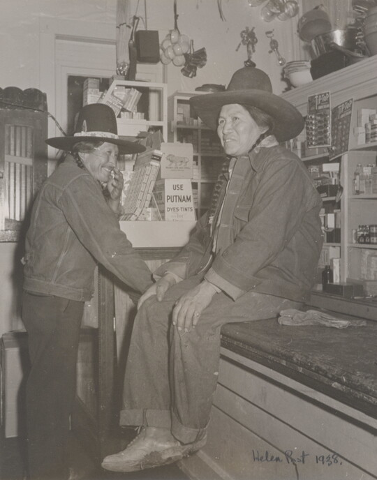 A black-and-white photograph of two Native American people in a general store: one wearing a jean jacket and cowboy hat sitting on a counter and the other dressed similarly standing nearby.