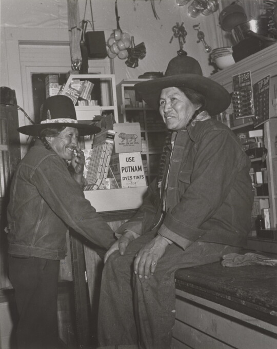 A black-and-white photograph of two Native American people in a general store: one wearing a jean jacket and cowboy hat sitting on a counter and the other dressed similarly standing nearby.