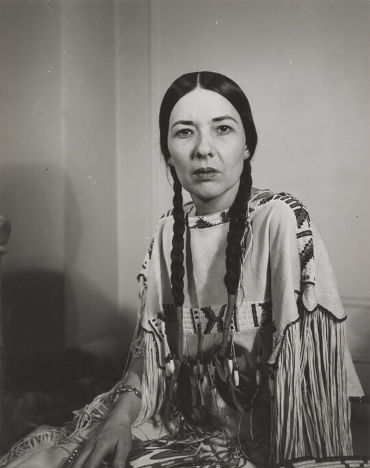 A black-and-white photograph of a seated Indigenous woman, hair in braids, gazing into the camera. 