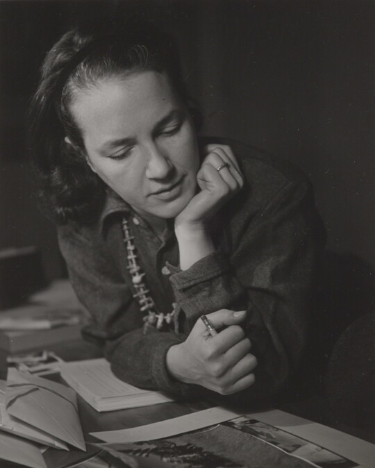 A black-and-white photograph of a White woman with her hand under her chin looking down at a photograph. 
