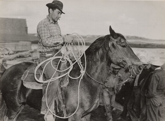 A black-and-white photograph of a light-skinned man in a brimmed hat holding a lasso on horseback.