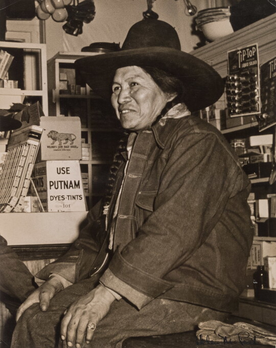 A black-and-white photograph of a Native American person wearing a jean jacket and cowboy hat sitting on a counter in a general store.