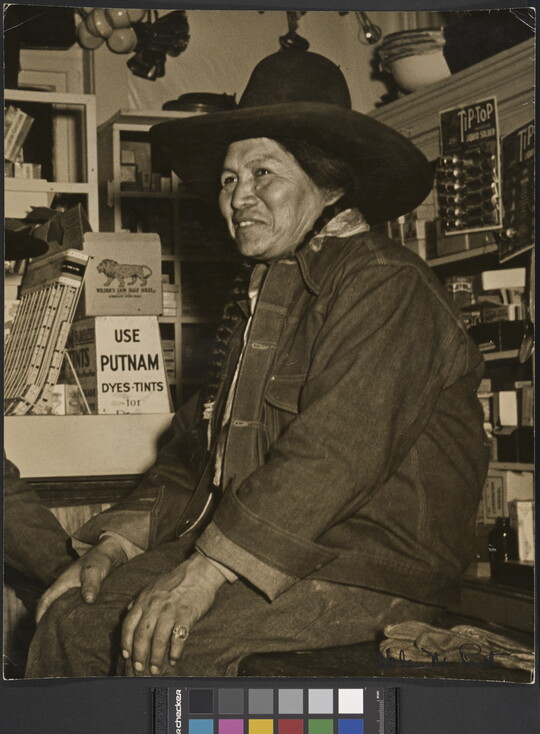 A black-and-white photograph of a Native American person wearing a jean jacket and cowboy hat sitting on a counter in a general store.