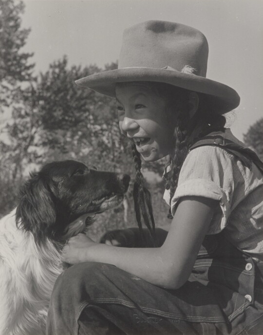 A black-and-white photograph of a smiling Native American child wearing a Homburg hat and sitting in front of a black-and-white dog.
