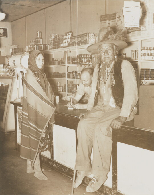 A sepia-toned photograph of a Indigenous man sitting on a counter, an Indigenous woman wrapped in a blanket standing at the counter, and a White man behind the counter. 