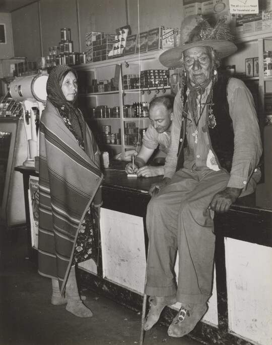 A black-and-white photograph of a Indigenous man sitting on a counter, an Indigenous woman wrapped in a blanket standing at the counter, and a White man behind the counter.