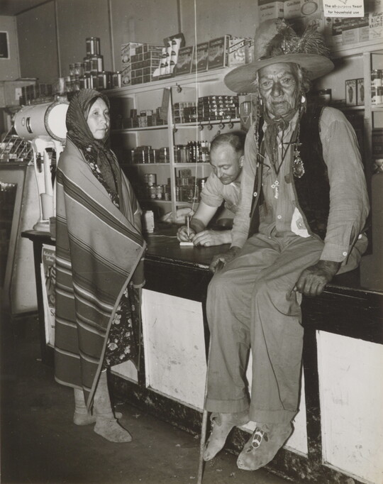 A black-and-white photograph of a Indigenous man sitting on a counter, an Indigenous woman wrapped in a blanket standing at the counter, and a White man behind the counter.