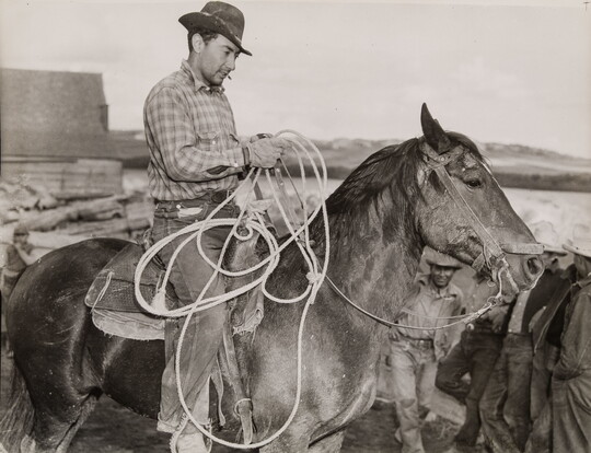 A black-and-white photograph of a light-skinned man in a brimmed hat holding a lasso on horseback.