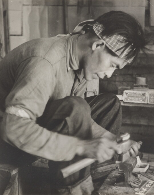 A black-and-white photograph of a seated Native American man bent over an anvil in a workshop.