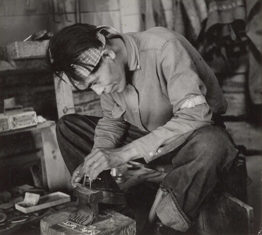 A black-and-white photograph of a seated Native American man bent over an anvil in a workshop.