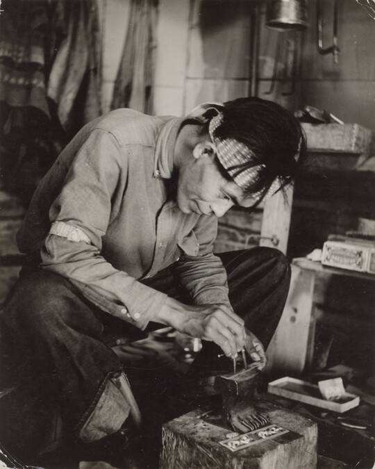 A black-and-white photograph of a seated Native American man bent over an anvil in a workshop.