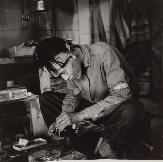 A black-and-white photograph of a seated Native American man bent over an anvil in a workshop.