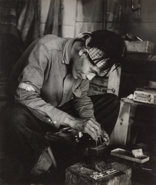 A black-and-white photograph of a seated Native American man bent over an anvil in a workshop.