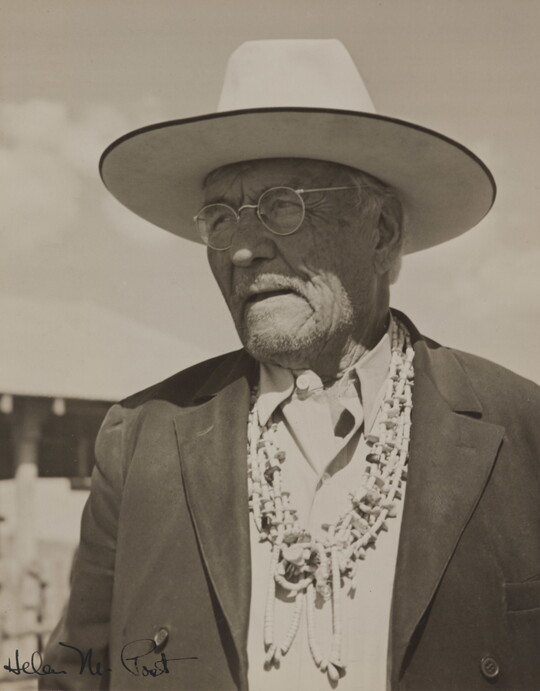 A black-and-white photograph of a Native American man wearing a wide-brimmed hat, glasses, a suit jacket and several necklaces.