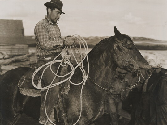 A black-and-white photograph of a light-skinned man in a brimmed hat holding a lasso on horseback.