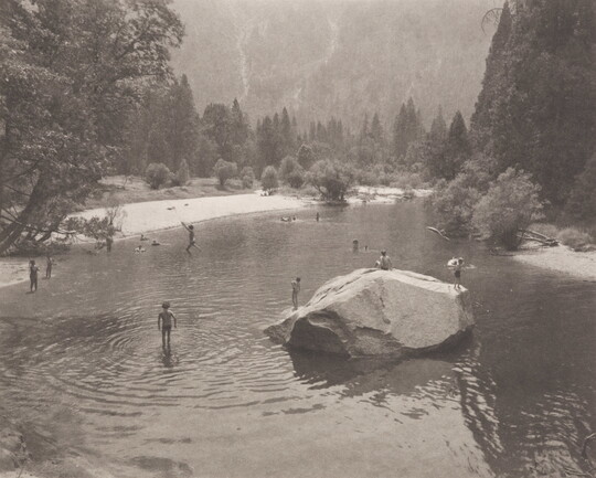 A black-and-white photograph of people swimming in a pond, a large stone in the center, surrounded by trees and mountains.