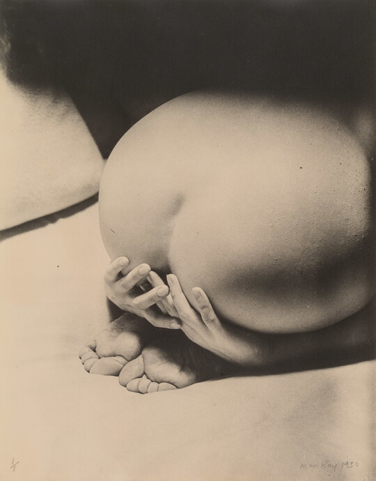 A black-and-white photograph of a White person from behind showing a bare bottom and back, feet tucked under, and hands partially covering their behind.