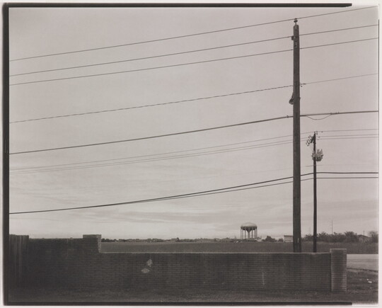 A black-and-white photograph of low brick wall beneath powerlines and utility poles and a water tower in the distance.