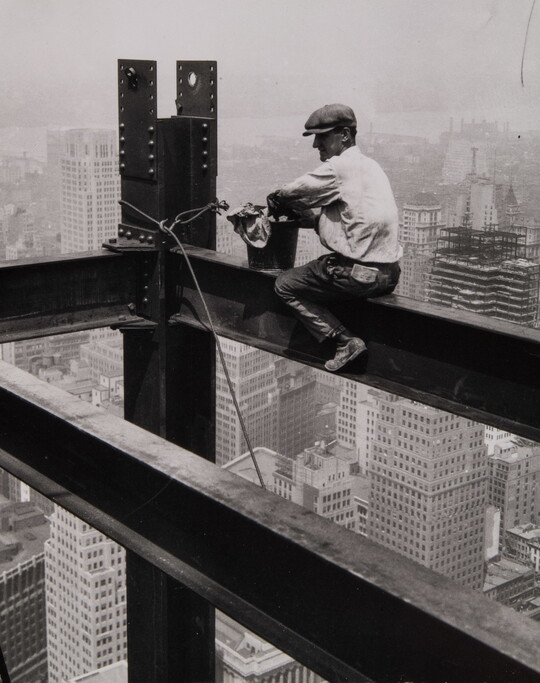 A black-and-white photograph of a man wearing a driving cap sitting on a steel beam high above a large city.