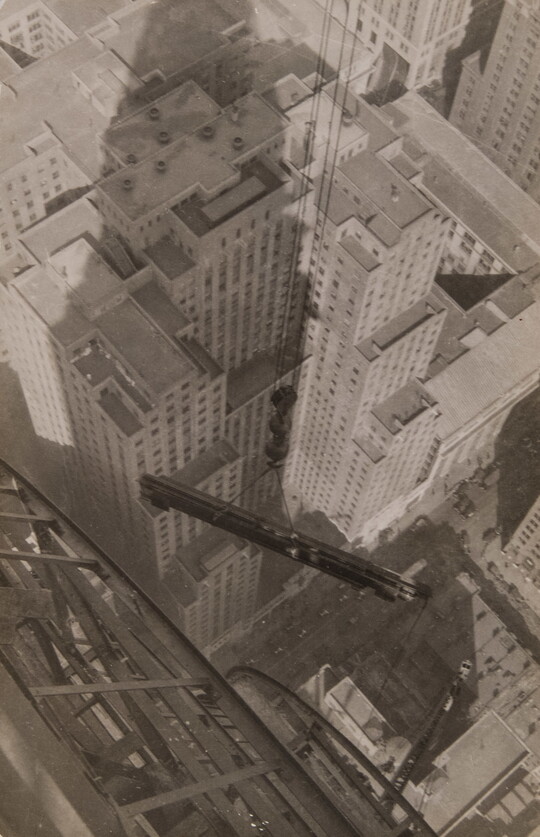 A black-and-white photograph looking from atop a skyscraper as a steel beam suspended by wire cables is lifted over a city.