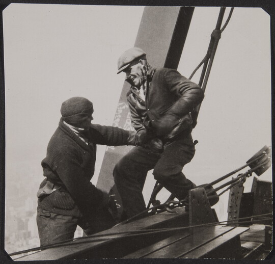 A black-and-white photograph of two men teasing one another as they balance on steel girders and cables high above the ground.
