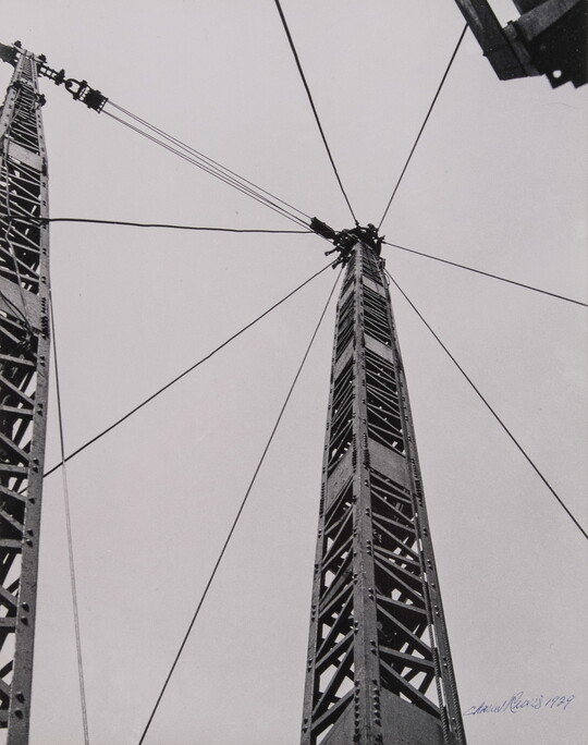 A black-and-white photograph of a steel derrick rising toward the sky with eight cables radiating from the top.