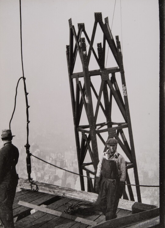 A black-and-white photograph of two men on a wooden platform of a construction site and a steel tower structure next to them.