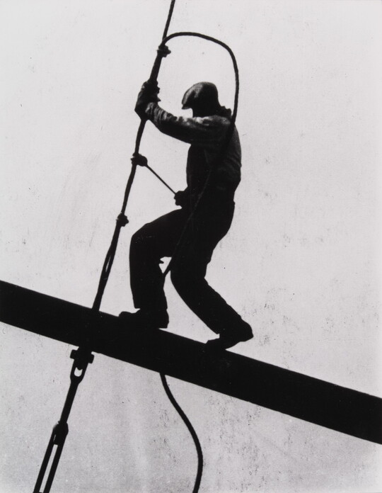 A black-and-white photograph of a man suspended precariously on an I-beam working with a tool on a cable, all in silhouette.