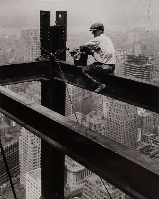 A black-and-white photograph of a man wearing a driving cap sitting on a steel beam high above a large city.