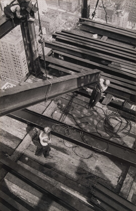 A black-and-white photograph from above of two men guiding a steel girder onto a platform high over a city.