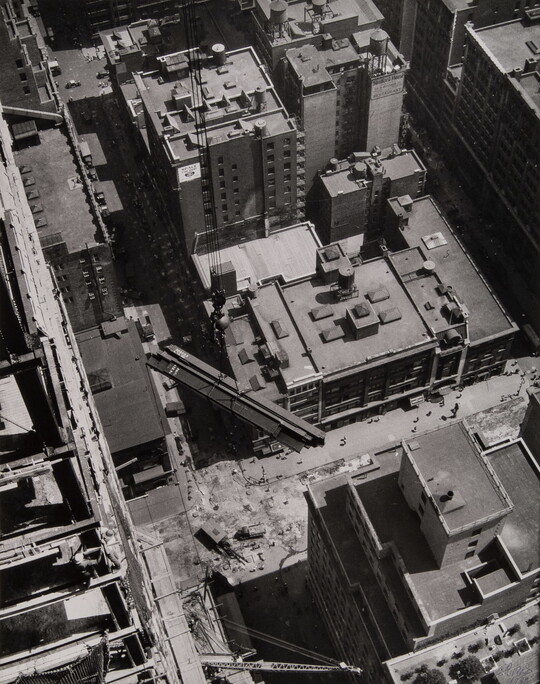A black-and-white photograph looking down on the buildings of a city and several steel girders suspended by cables in mid-air.
