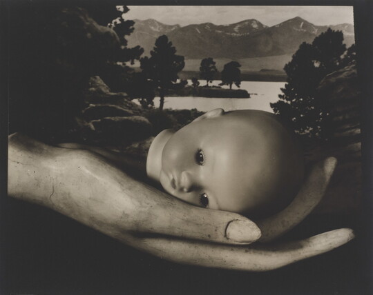 A black-and-white photograph of a mannequin hand holding a baby doll head in front of a landscape backdrop.