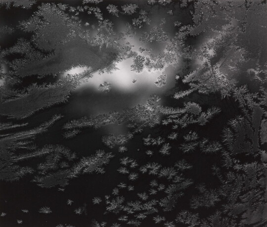 An up-close black-and-white photograph of frost gathering on a clear surface.