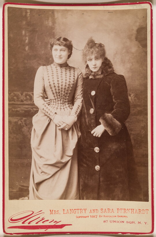 A sepia-toned studio portrait photograph of two elegantly dressed women in 1880s clothing above a bold red signature by the photographer.
