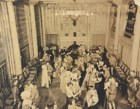 A sepia-toned photograph of a large group of people dancing in a large room, as viewed from above.
