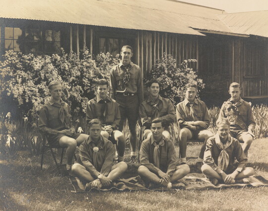 A sepia-toned photograph of a group of light-skinned young men in uniforms posed outside a building.