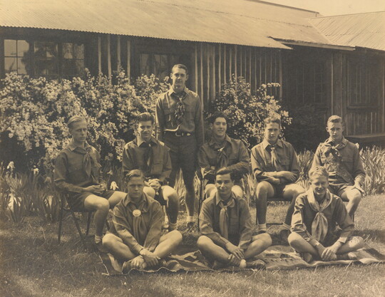 A sepia-toned photograph of a group of light-skinned young men in uniforms posed outside a building.