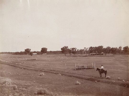 A black-and-white photograph of a person on horseback in a grassy field with trees in the background.