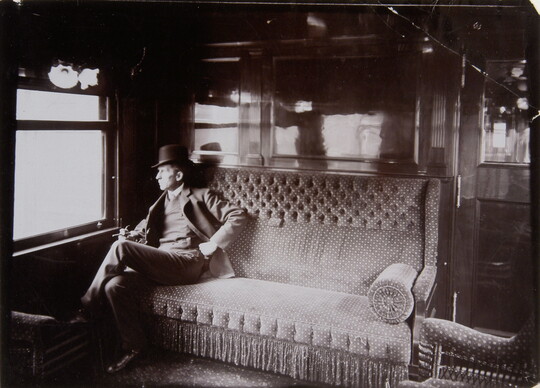 A black-and-white photograph of a White man wearing a top hat sitting on a couch in a railroad car, gazing out the window.