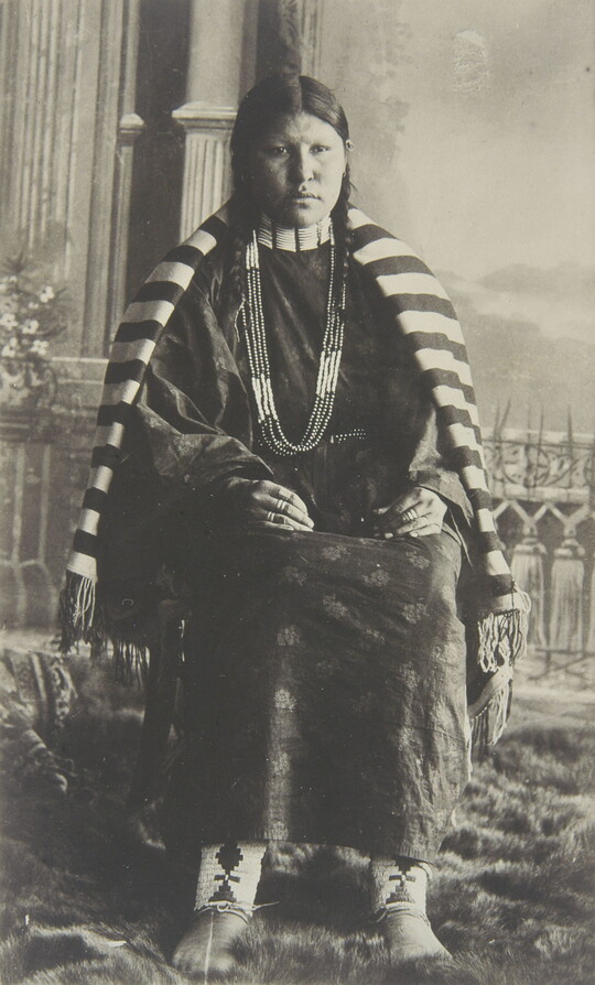 A black-and-white photograph of a seated Native American woman in traditional clothing with a striped blanket over her shoulders.