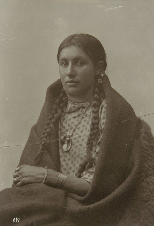 A black-and-white photograph of a seated Native American woman in traditional clothing with her hair in braids and a blanket around her shoulders.