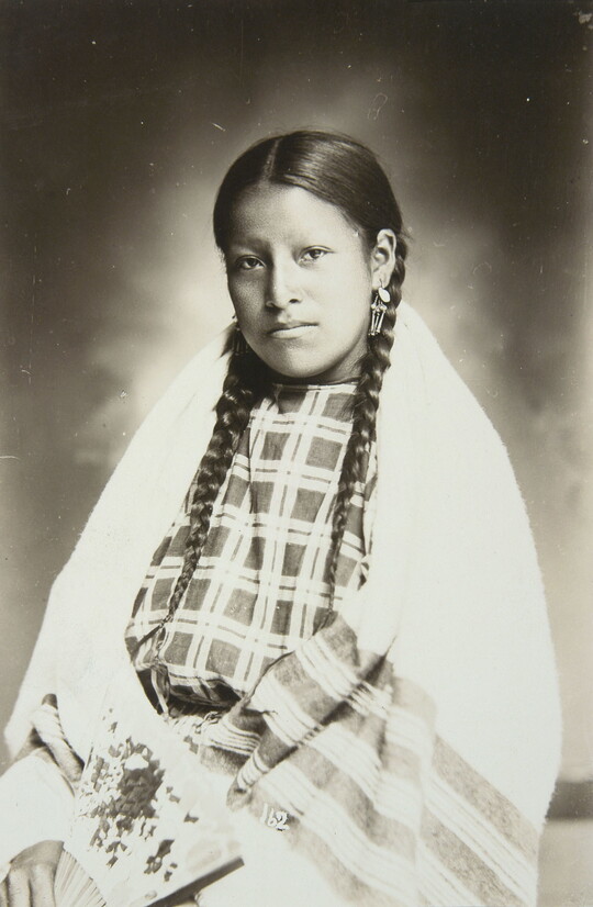 A formal black-and-white portrait photograph of a seated Native American girl, hair in braids, wearing cultural dress, and holding a fan.