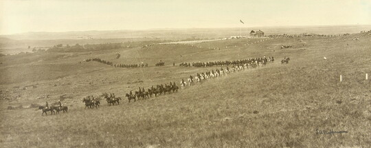 A black-and-white photograph of a grassy plain with a line of cavalry on horseback travelling through the landscape.