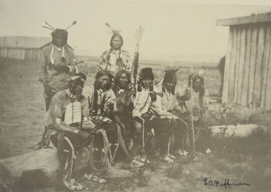 A black-and-white photograph of six Native Americans sitting on a log and two standing behind them, all wearing traditional clothing.