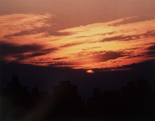 A color photograph of an orange and purple sky during a sunrise, and the dim silhouette of a city skyline.