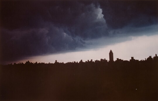 A color photograph of dark blue-gray storm clouds over the silhouette of a city skyline.