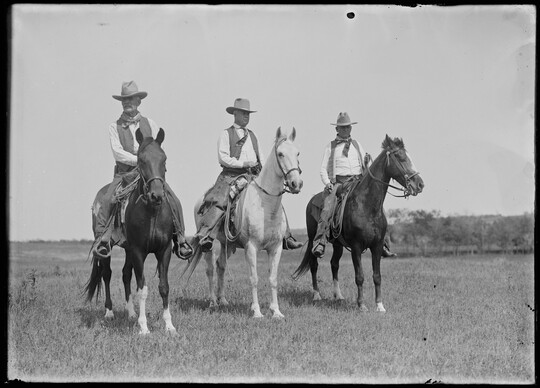 A black-and-white image of three White cowboys on horseback in a grassy field.