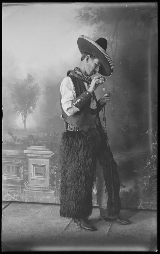 A black-and-white, studio portrait image of a man standing in front of a backdrop wearing a sombrero, a kerchief around his neck, a vest, animal fur chaps, and cowboy boots.