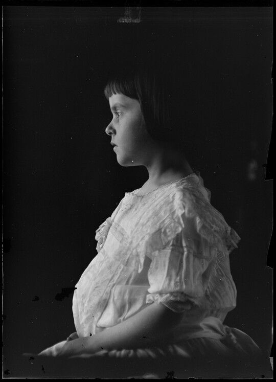A black-and-white image of a young White girl in profile wearing a white dress.