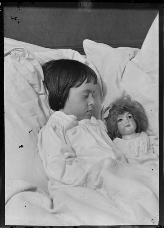 A black-and-white image of a young White girl, surrounded by pillows, sleeping next to a doll.